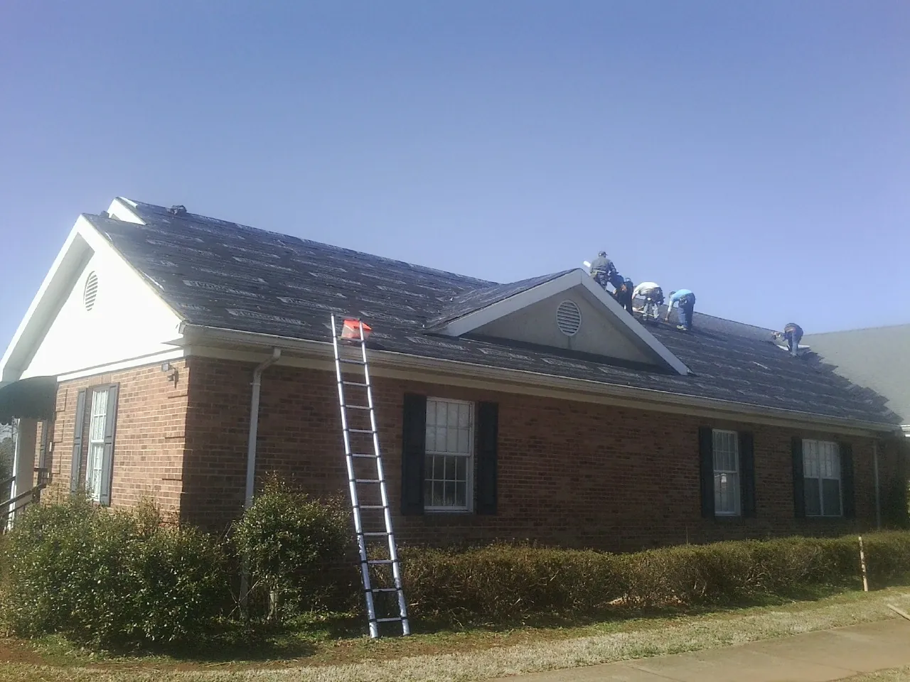 Crew tearing shingles off a brick ranch roof with a ladder set — Metal Roof Repair work by our crew in Gilman