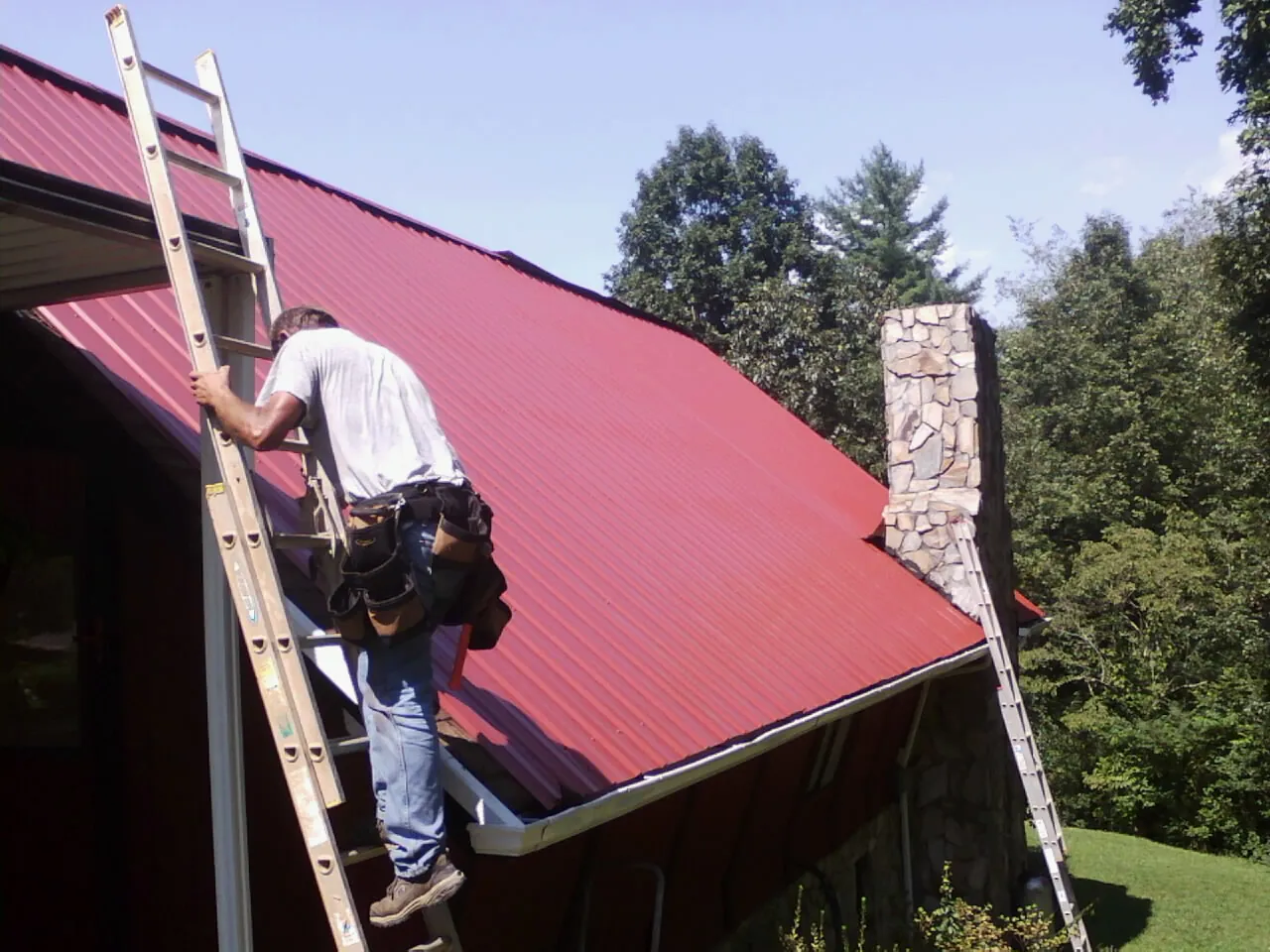 Roofer on a ladder beside a red metal roof and stone chimney — Soffit & Fascia Repair by Roofing Brothers in Gilman